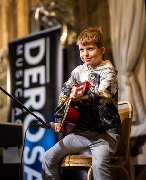Young boy holding a guitar in front of a microphone and 'Dero' sign.